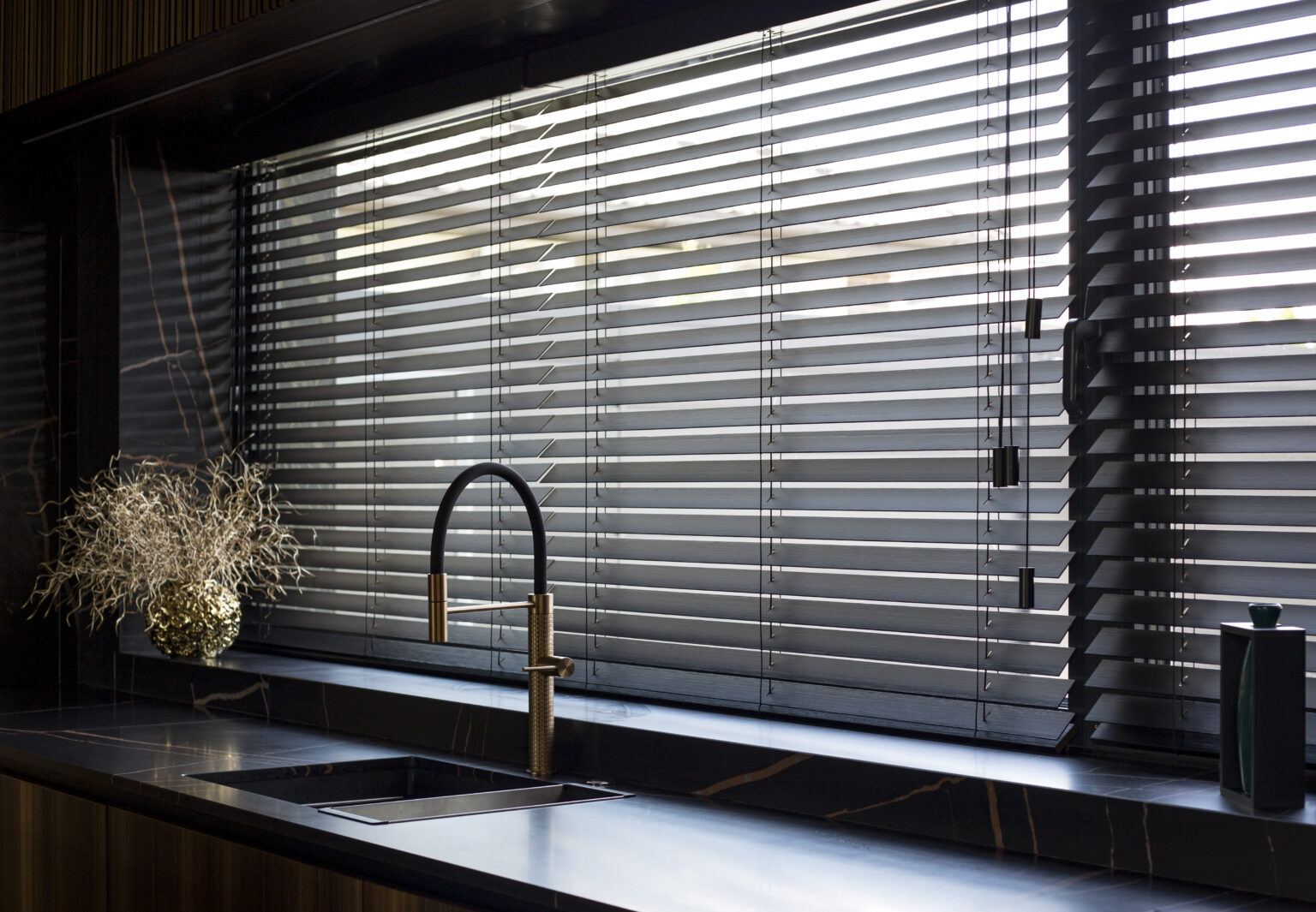 Wooden blinds black color closeup on the window. Bamboo slats 50mm wide. Venetian wood blinds in the kitchen. Black tapes. Sink with copper faucet near the window. Round vase is on the windowsill.