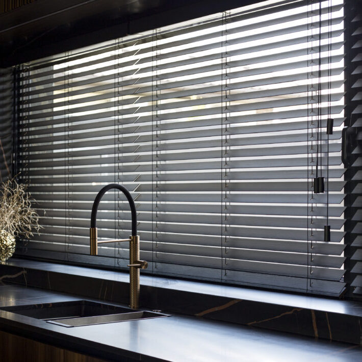 Wooden blinds black color closeup on the window. Bamboo slats 50mm wide. Venetian wood blinds in the kitchen. Black tapes. Sink with copper faucet near the window. Round vase is on the windowsill.